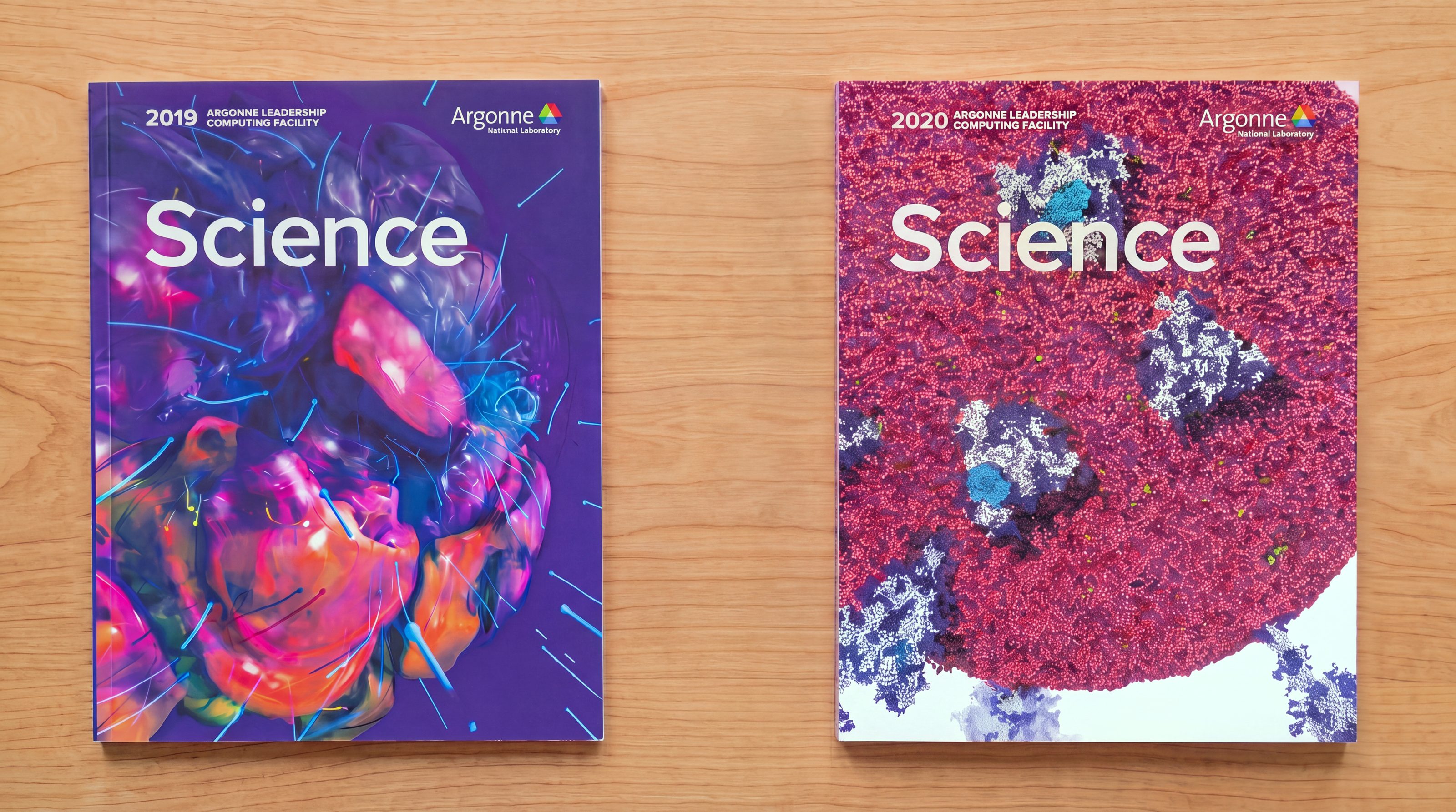 Two printed ALCF Science reports side by side on a wood table: the 2019 cover carrying a purple and orange fluid simulation threaded with cyan rods, the 2020 cover a dense field of red particles with pale clusters running through it.