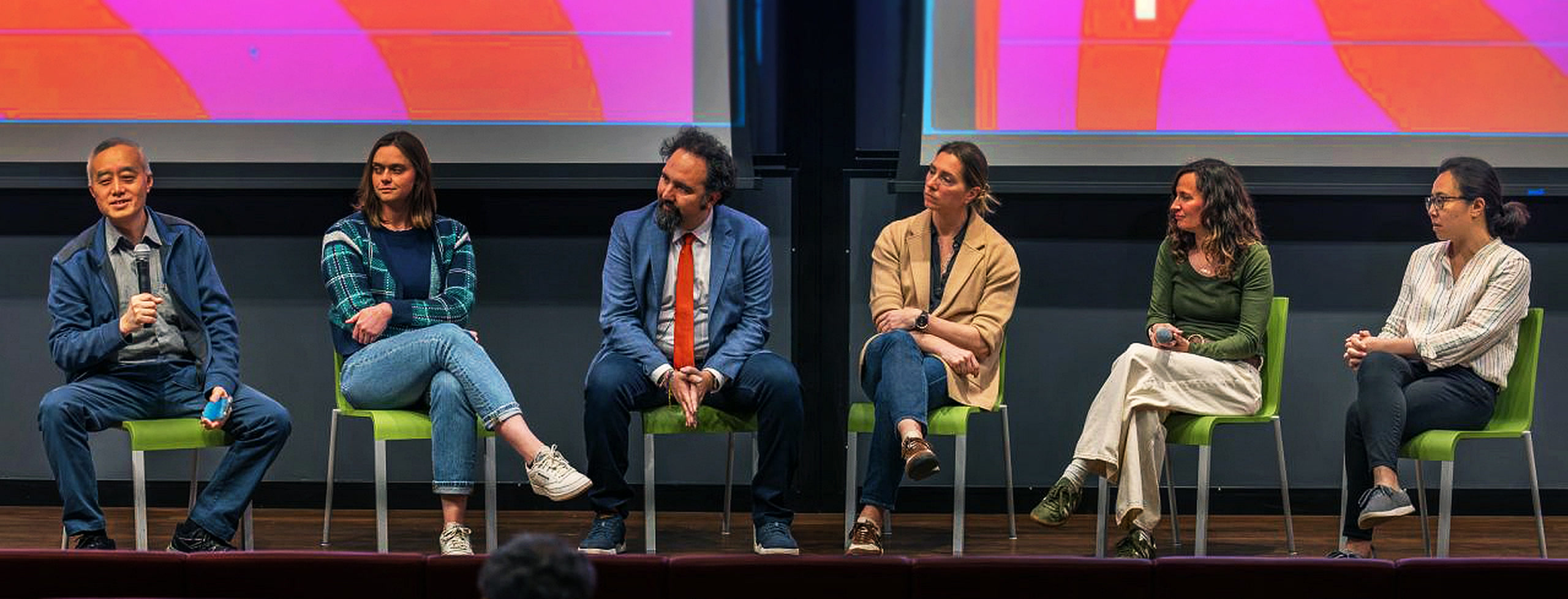 A Fusion Energy Week panel discussion: six speakers seated on green chairs on a stage, one speaking into a microphone, beneath two screens showing the week&rsquo;s pink and orange graphics.
