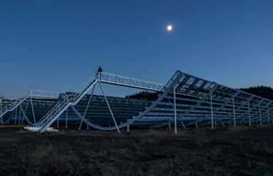 The CHIME telescope's long half-pipe reflectors under a deep blue twilight sky, the moon above them.
