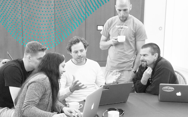 Collaboration members around a table of laptops, mid-conversation, the NANOGrav dot pattern on the wall behind them.