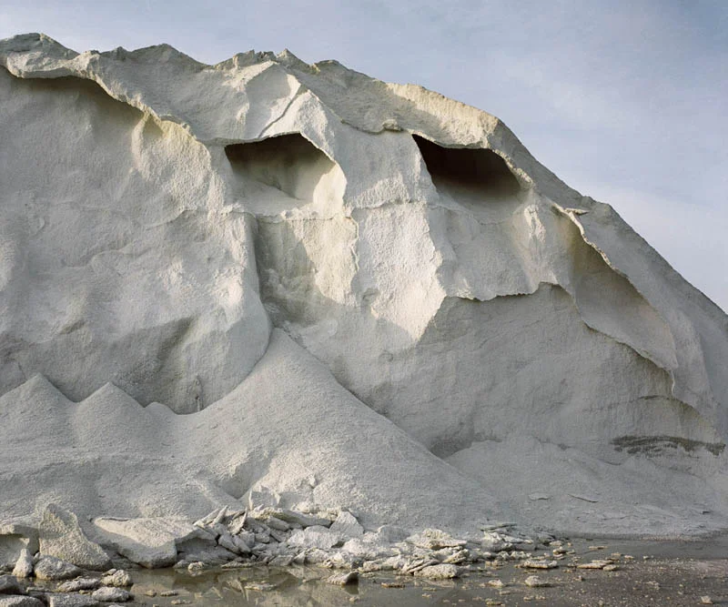 A towering white salt mound eroded into sharp ridges and hollow ledges, above a shallow puddle scattered with broken crust.