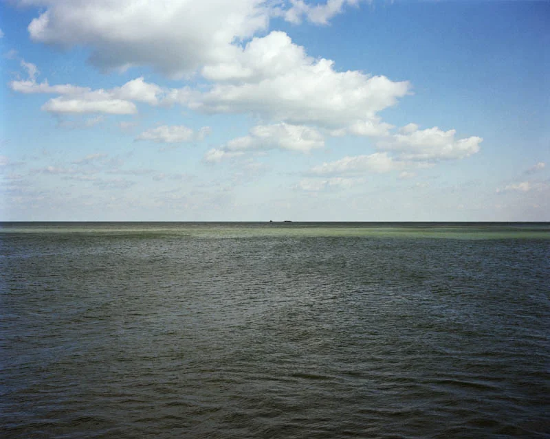 Green-gray water meets a flat horizon with a distant boat, under a blue sky of scattered white clouds.
