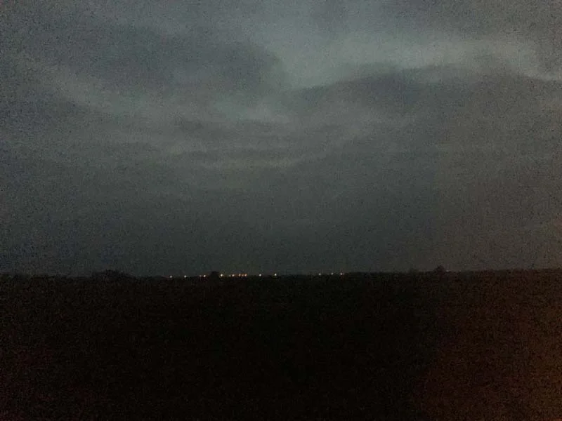 A dark, grainy night sky of low storm clouds over a black field, with a thin row of distant lights on the horizon.