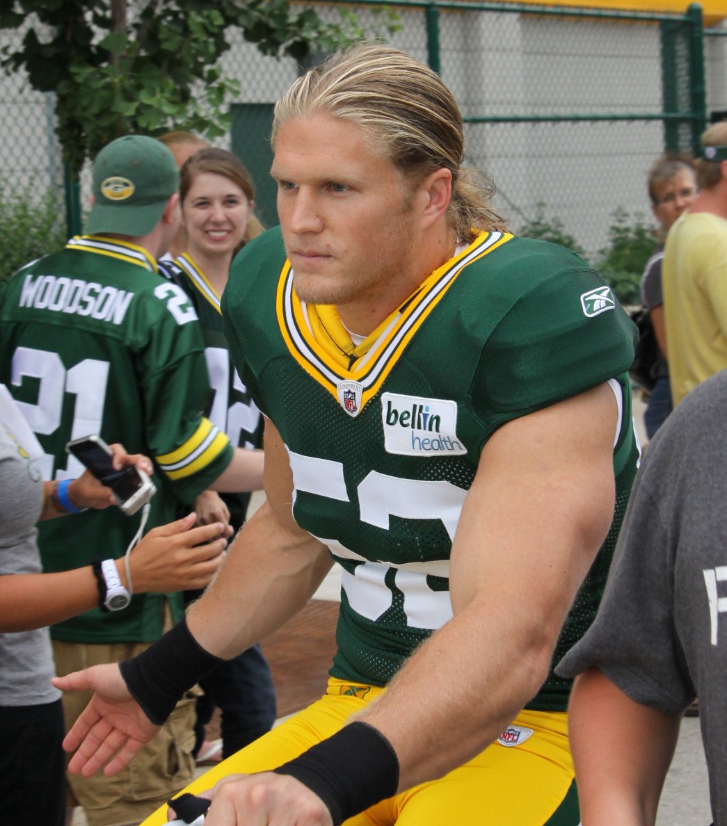 A long-haired blond football player in a green and gold practice jersey passes a crowd of fans along a chain-link fence.