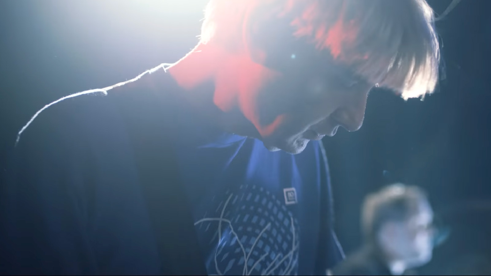 A musician in a blue t-shirt bows their head in a bright stage light streaked red, a bandmate blurred behind.